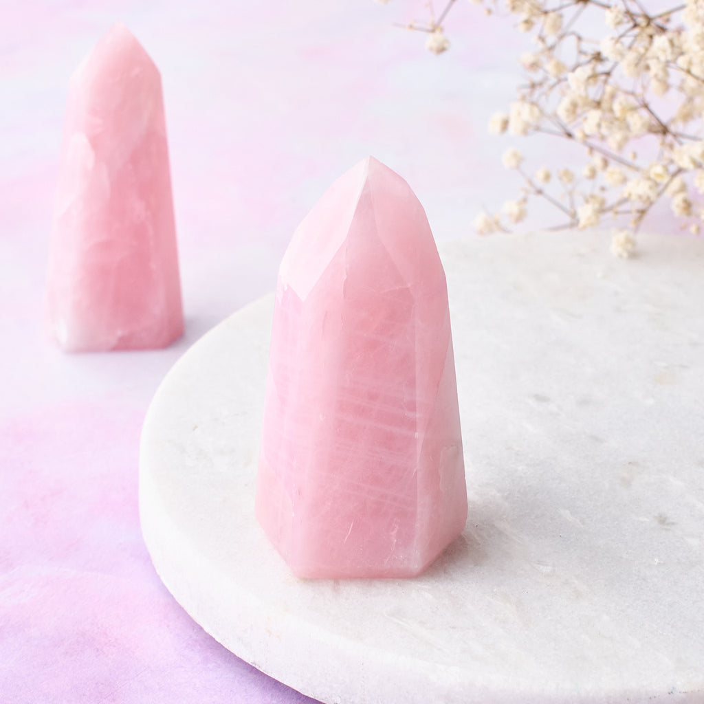 En närbild framifrån på ett litet rosenkvartstorn med ytterligare ett litet rosenkvartstorn synligt i bakgrunden, mot en vit bakgrund med rosa-lila toner.