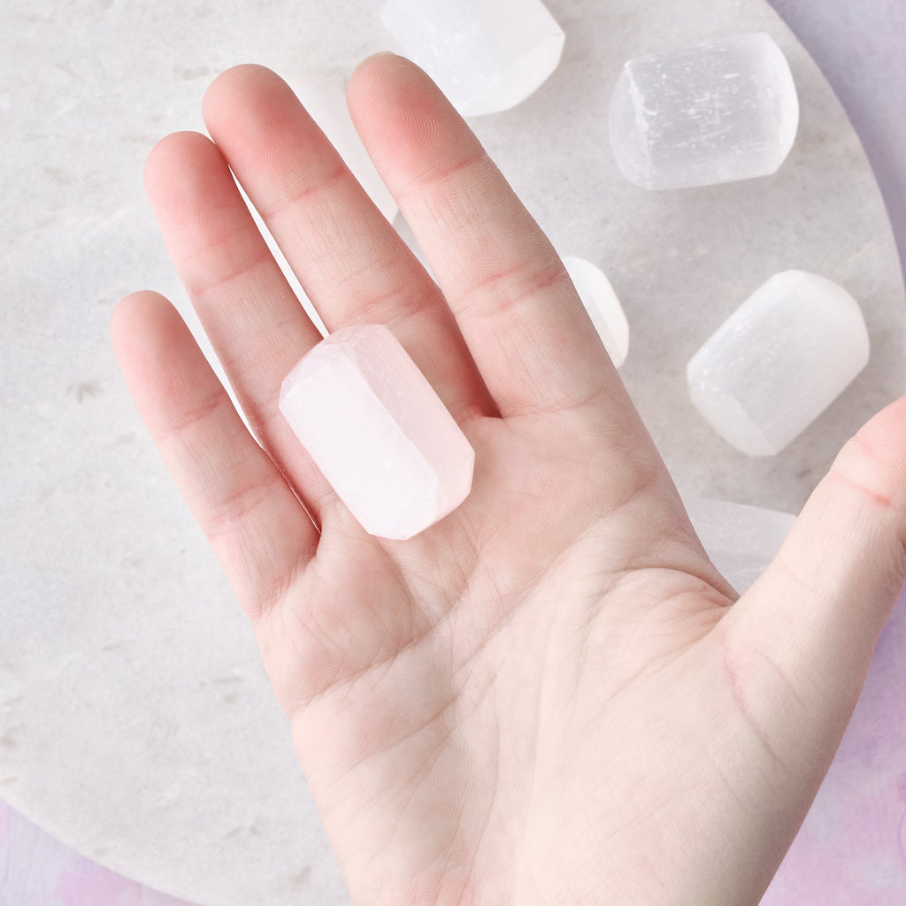 En liten trumlad selenit sedd uppifrån när den hålls i en hand, mot en vit bakgrund med rosa-lila toner.