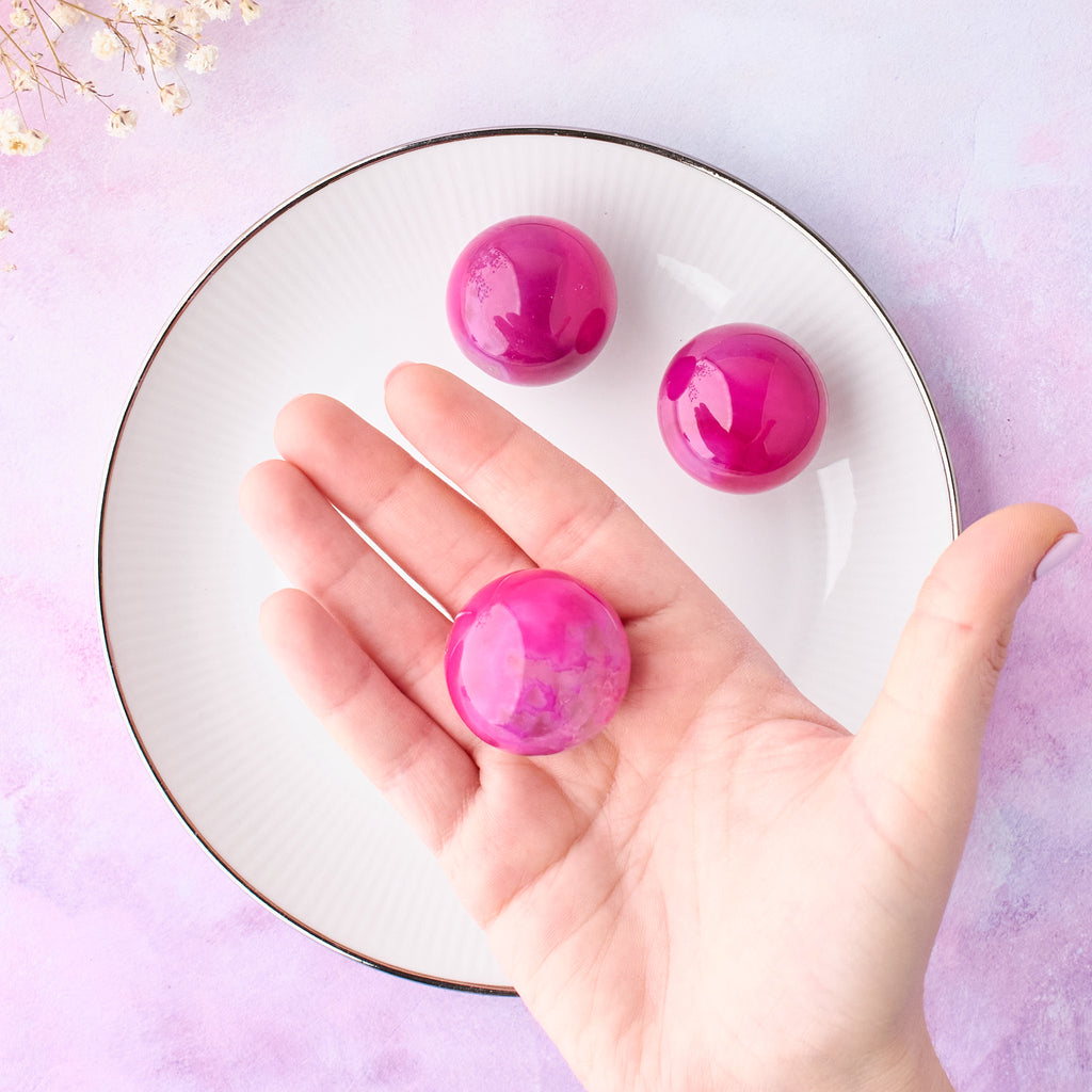 Tre rosa agat klot, ett i handen och två på vit tallrik, fotograferade uppifrån med rosa-lila bakgrund